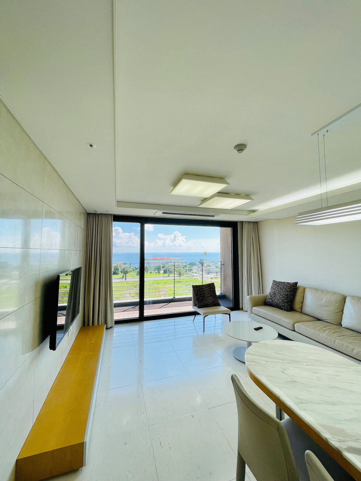 A bright living area features a sofa and two upholstered chairs arranged around a circular coffee table. Large windows frame an ocean view, allowing natural light to fill the space. Light fixtures are mounted on the ceiling, complementing the neutral decor.