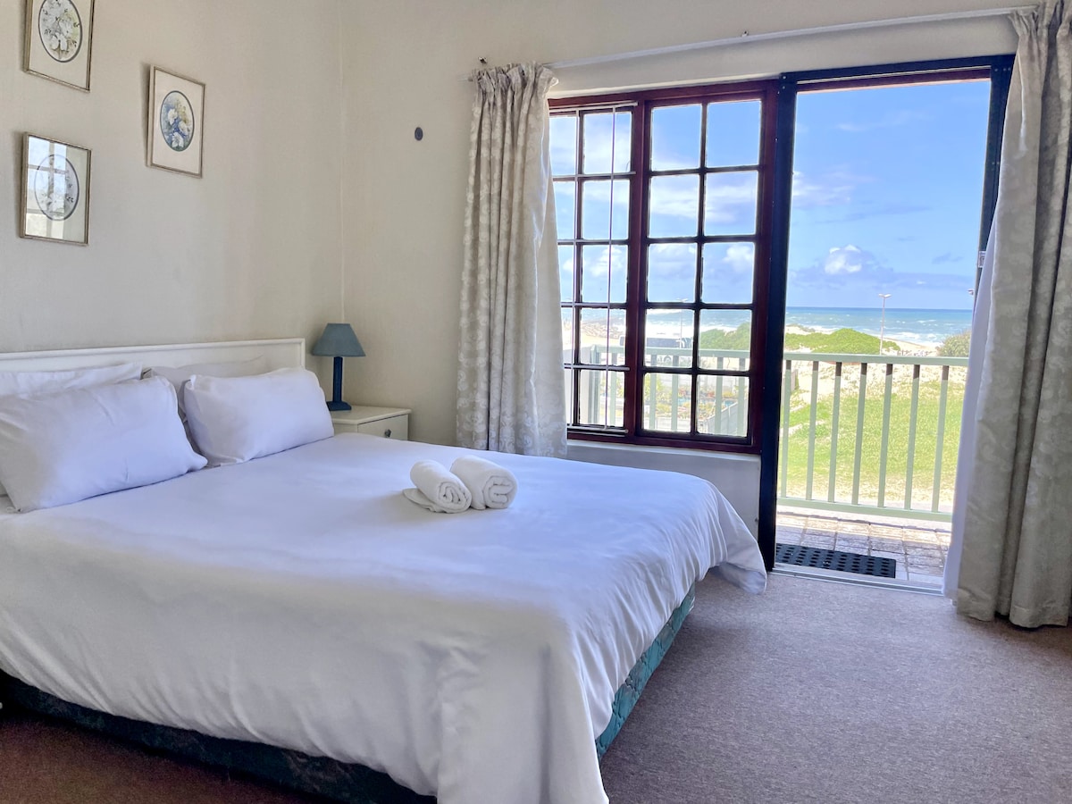 A bright bedroom features a spacious bed with freshly laundered linens and neatly rolled towels placed at the foot. Natural light filters in through large doors, leading to a balcony that offers a view of the outdoors and the nearby beach.