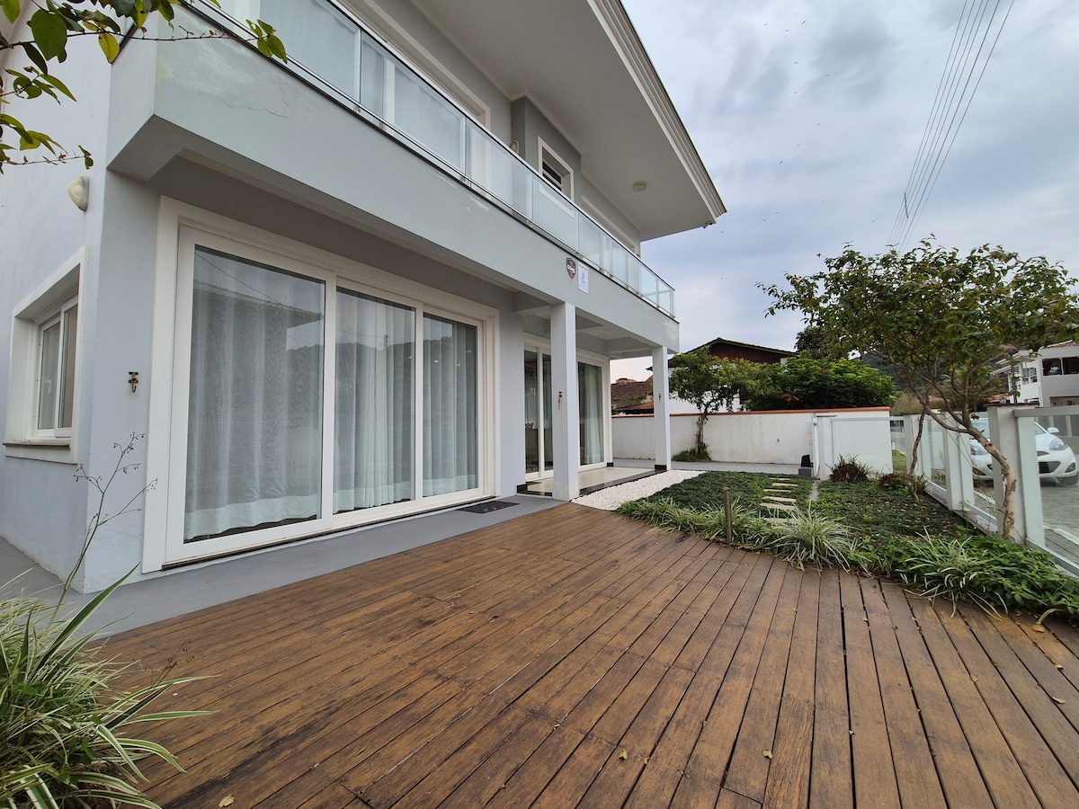 A spacious outdoor deck features polished wooden flooring, surrounded by greenery. Large glass doors provide access to the interior, allowing natural light to enhance the area. The setting is complemented by a well-maintained garden with neatly trimmed plants.