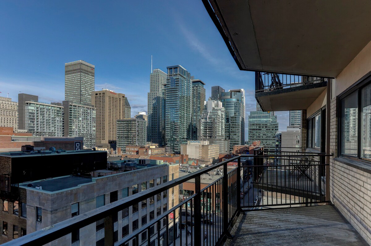 A private balcony offers a view of the downtown Montreal skyline, showcasing a mix of modern skyscrapers and historic buildings. The balcony features a simple railing and concrete flooring, providing a space for outdoor relaxation and city observation.