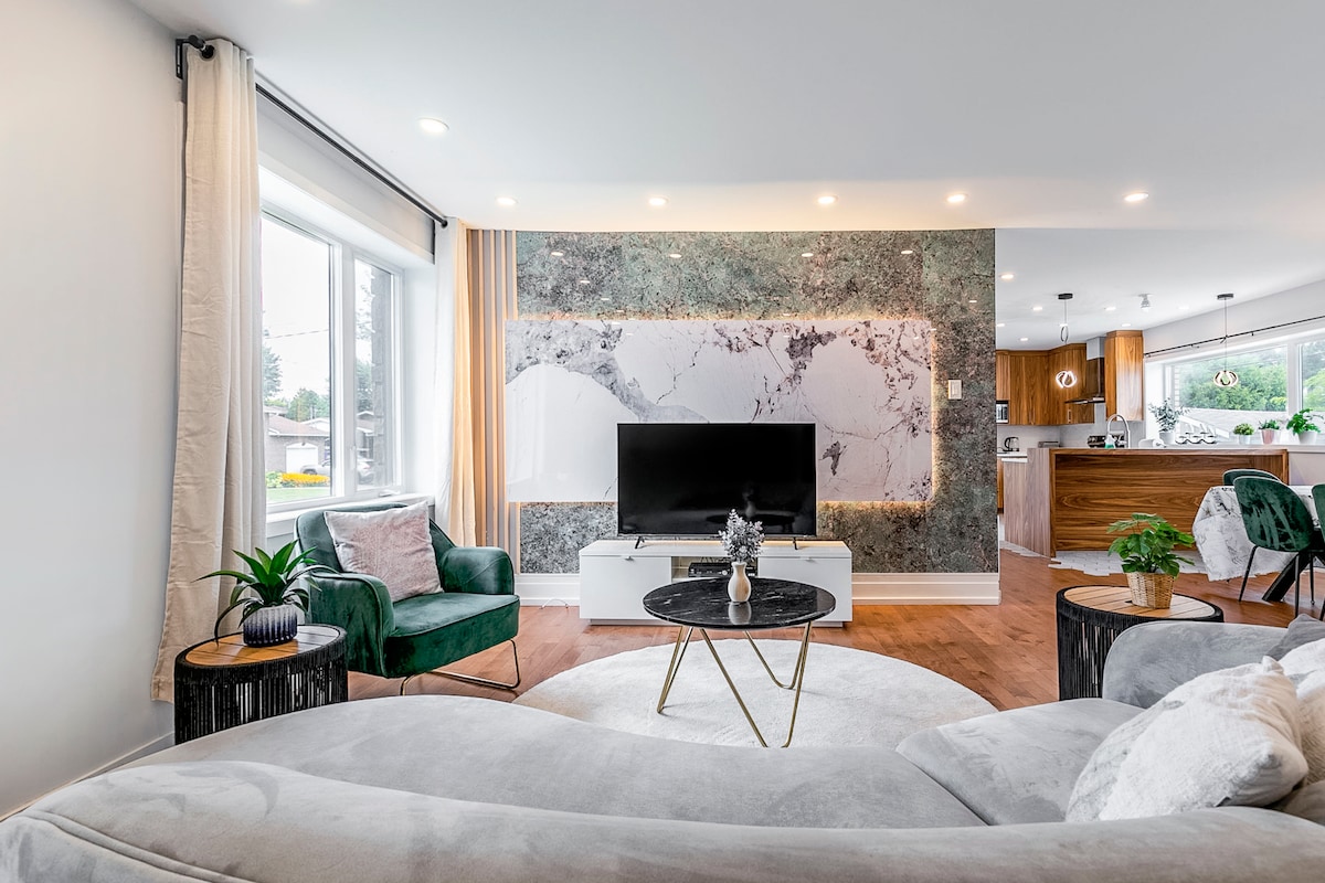 A modern living area features a large television on a sleek white console, with an elegant marble accent wall behind it. A cozy gray sofa is paired with green accent chairs, and natural light filters through a nearby window, highlighting the wooden flooring.