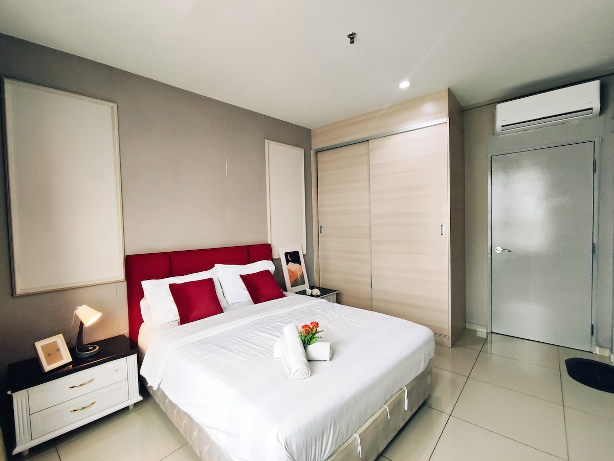 A neatly arranged sleeping space features a spacious bed with red accent pillows. A decorative arrangement of fresh flowers sits atop the bed. A wardrobe with sliding doors is visible, along with a bedside lamp and a small framed artwork on the nightstand.