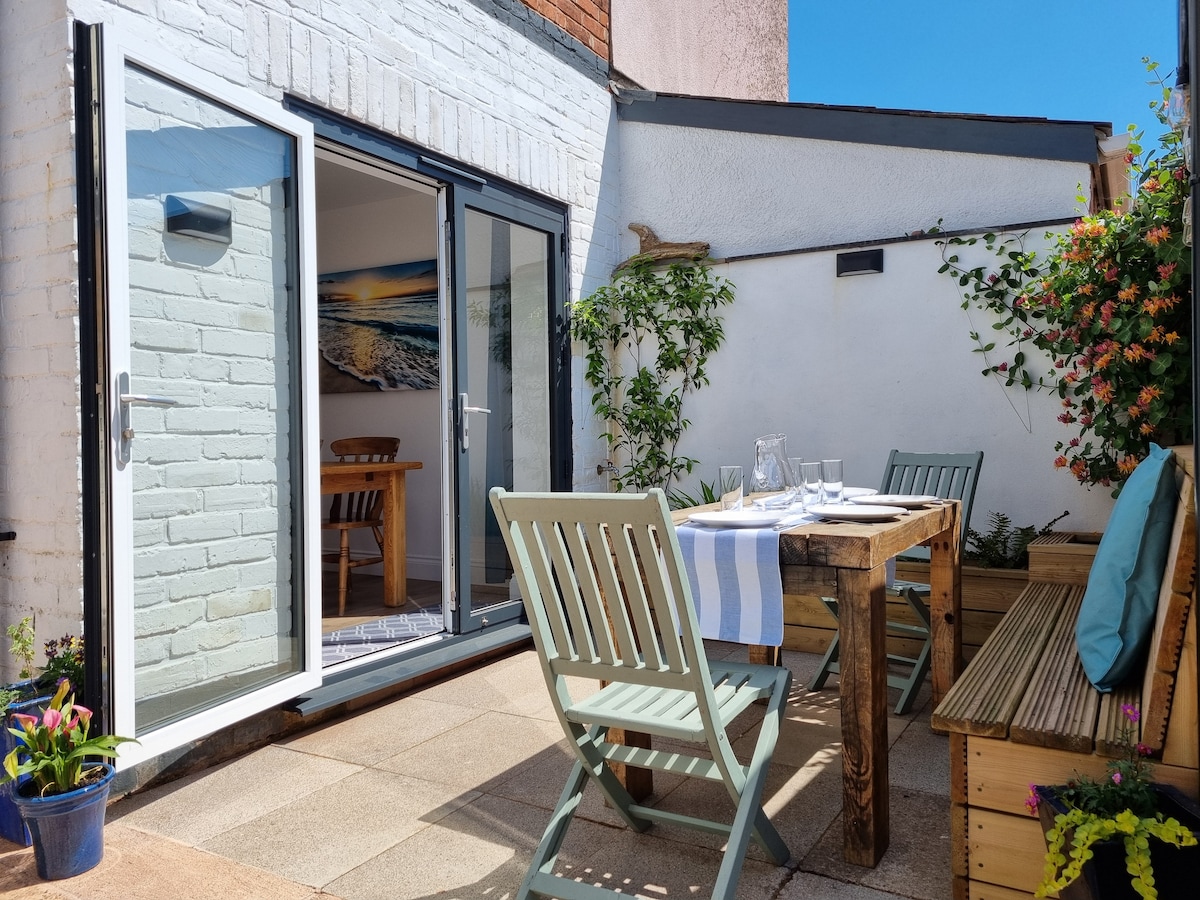 A private courtyard garden features a wooden dining table set for outdoor meals. Green chairs surround the table, while climbing plants and flowers enhance the setting. Large double doors open into the cottage, allowing natural light to fill the space.