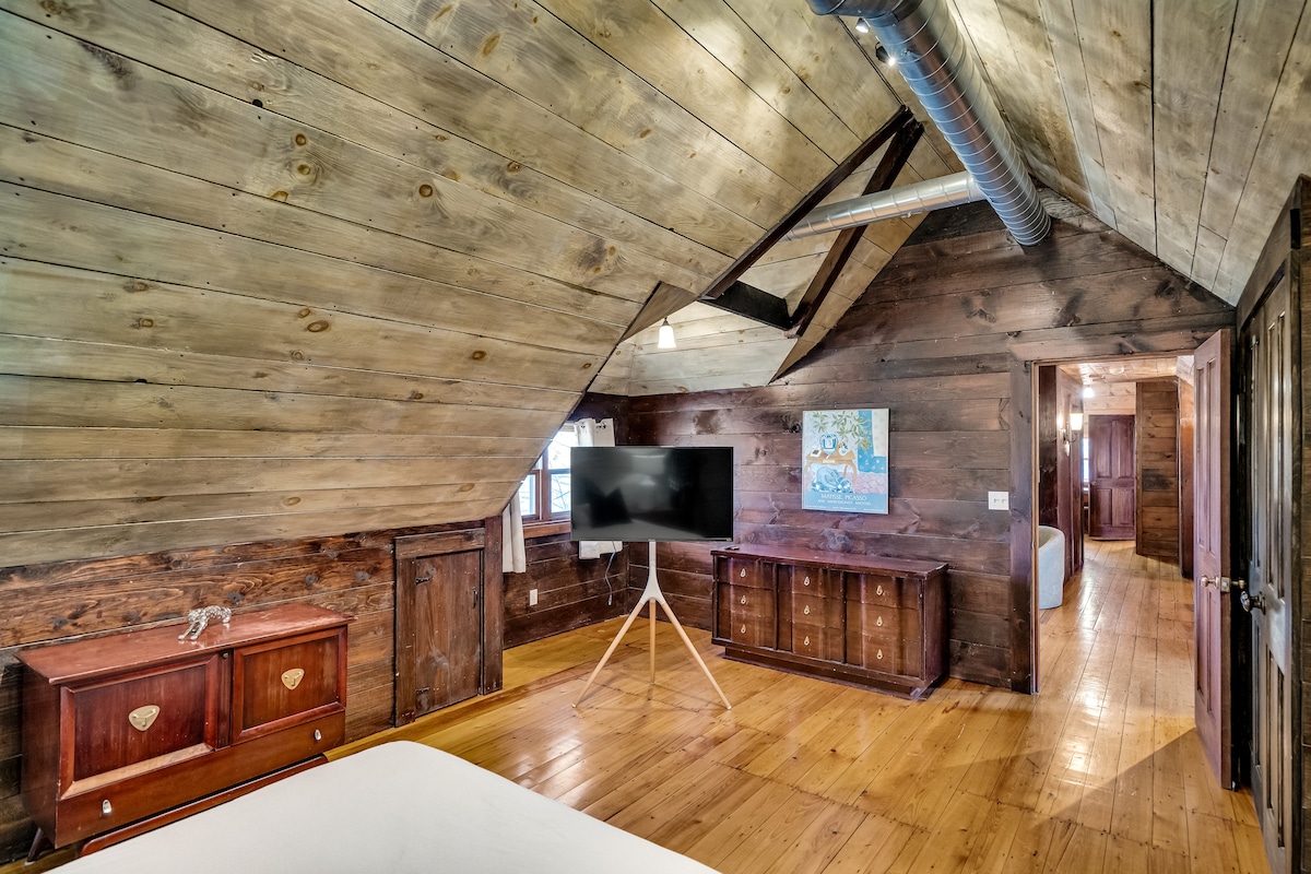 The spacious master bedroom features rustic wooden walls and a sloped ceiling, creating a cozy ambiance. A simple white bed is positioned adjacent to a sleek TV mounted on a stand. Natural light streams in through multiple windows, illuminating polished wooden floors.