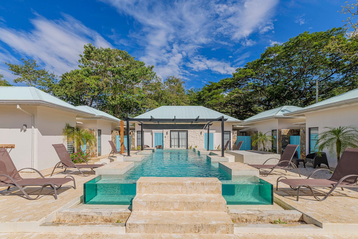 A serene outdoor space features a central swimming pool surrounded by elegant loungers. The clear water reflects the blue sky above, complemented by tropical greenery in the background. Two villas frame the pool area, enhancing the inviting atmosphere.