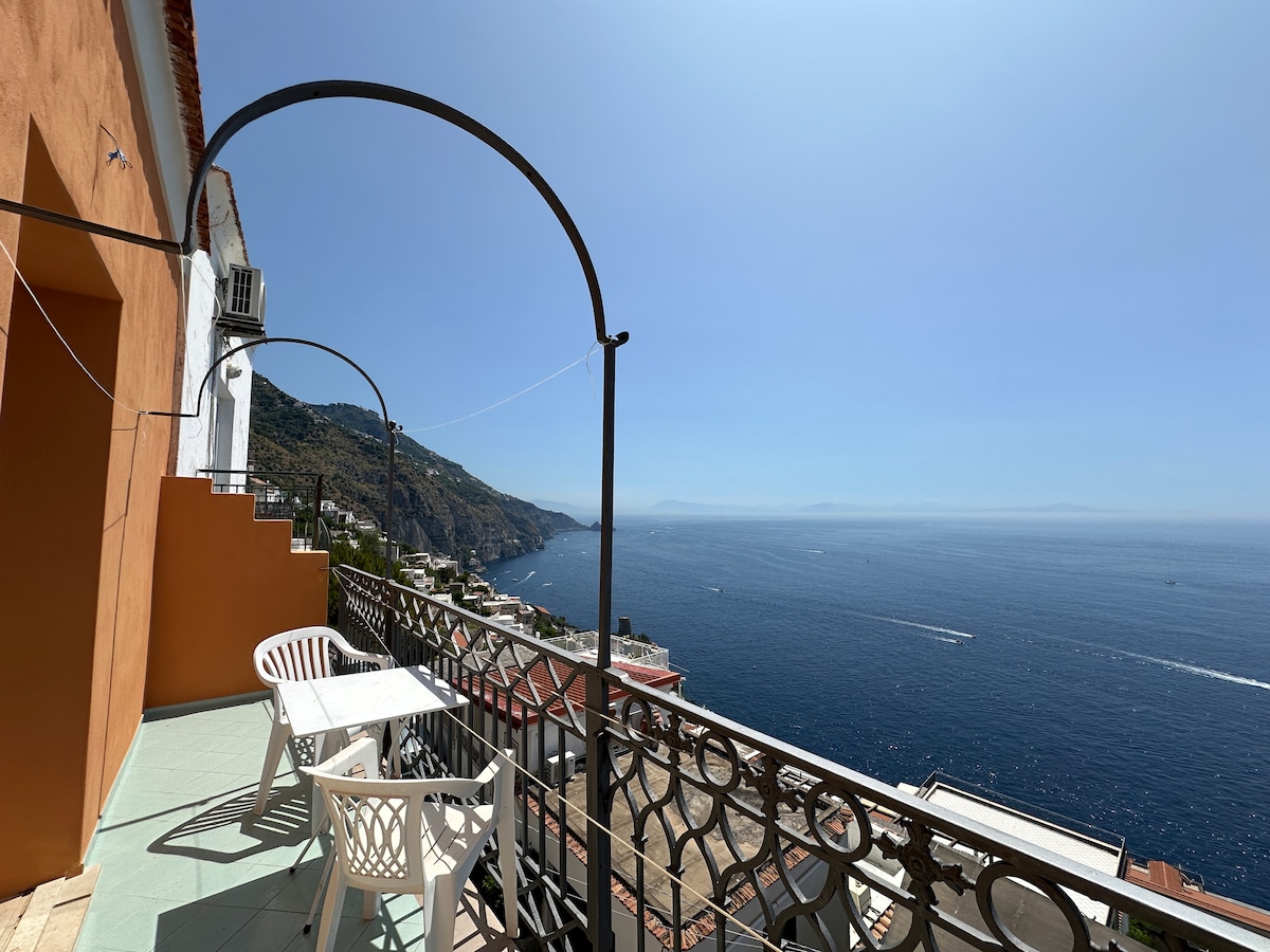 A sunlit balcony offers panoramic views of the sea and coastal landscape. Two white chairs are positioned on a tiled surface, complemented by a protective railing. The clear blue sky merges with the water in the distance, enhancing the sense of openness.