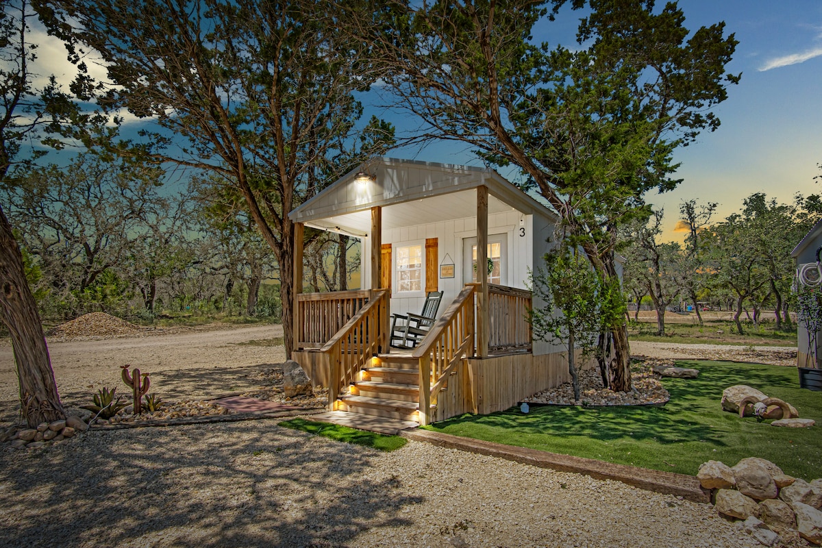 A charming cabin entrance is highlighted by a covered porch with rocking chairs. Surrounding natural greenery and soft outdoor lighting create an inviting pathway leading to the door, emphasizing a connection with the tranquil outdoors.
