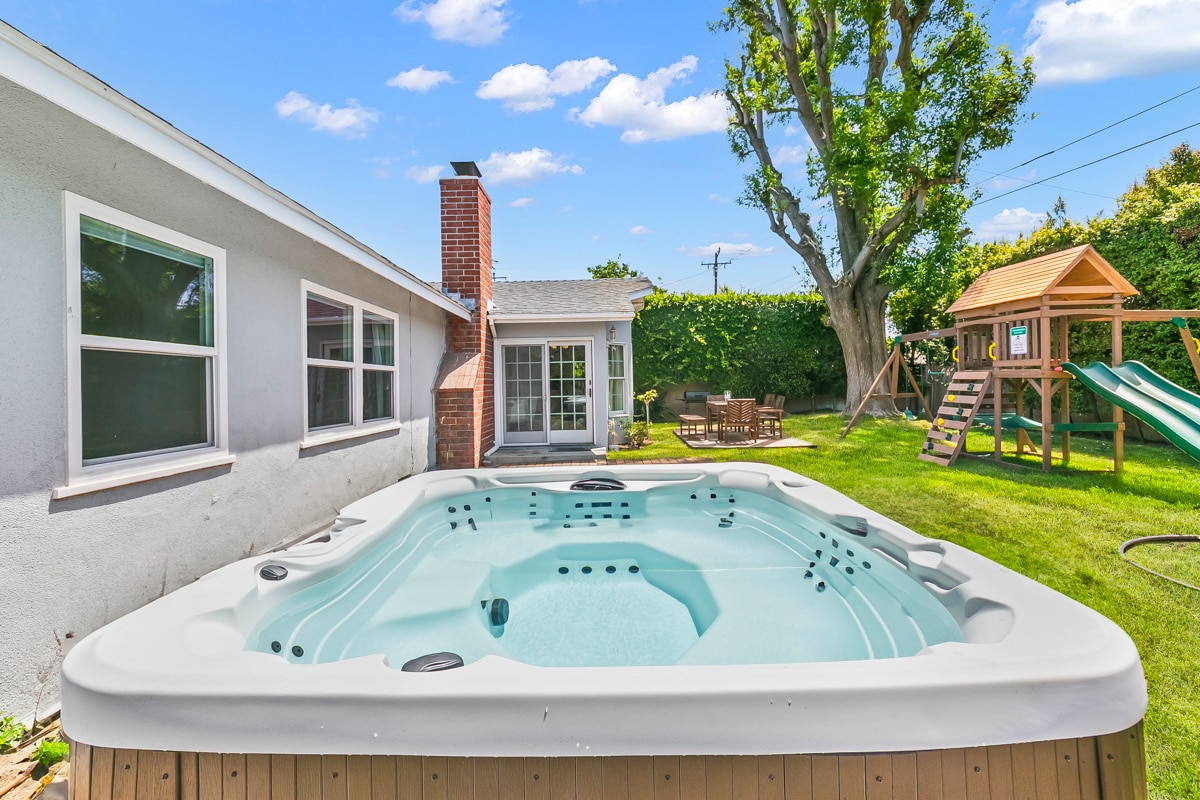 A well-maintained backyard features a jacuzzi surrounded by a spacious deck. Nearby, a playground is visible, offering play structures and slides. A green lawn extends further, with seating available within view of the house's large window, allowing for outdoor relaxation and enjoyment.