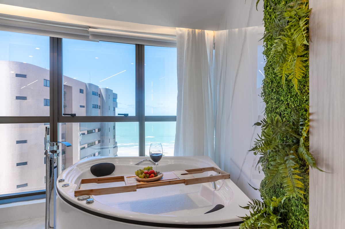 A private hot tub is positioned by a large window, offering a view of the ocean. A wooden tray holds a glass and fresh fruit, while greenery adds a touch of nature to the space. Light filters through sheer curtains, enhancing the relaxing atmosphere.