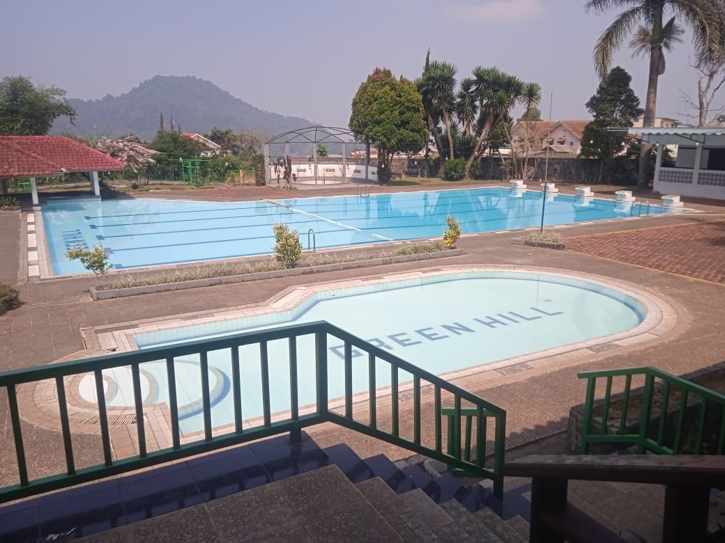 An outdoor pool area is showcased, featuring several swimming pools surrounded by well-maintained gardens. A small pool labeled 'GREEN HILL' sits next to a larger swimming pool, with a view of distant hills in the background. Sunlight reflects off the water's surface.