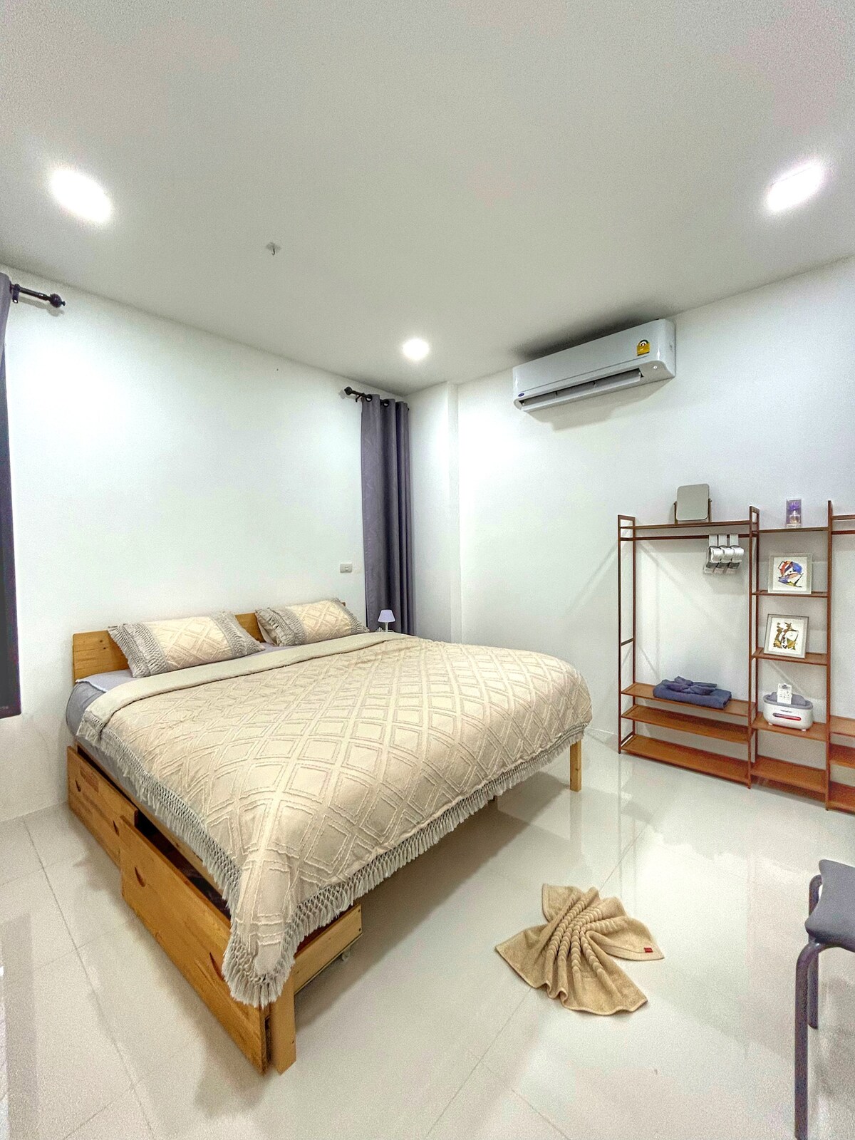 A spacious room features a wooden bed with a patterned blanket, complemented by matching pillows. Soft lighting from ceiling fixtures illuminates the area, while a decorative shelf displays toiletries and folded towels. A plush rug is positioned near the bed, enhancing the room's comfort.