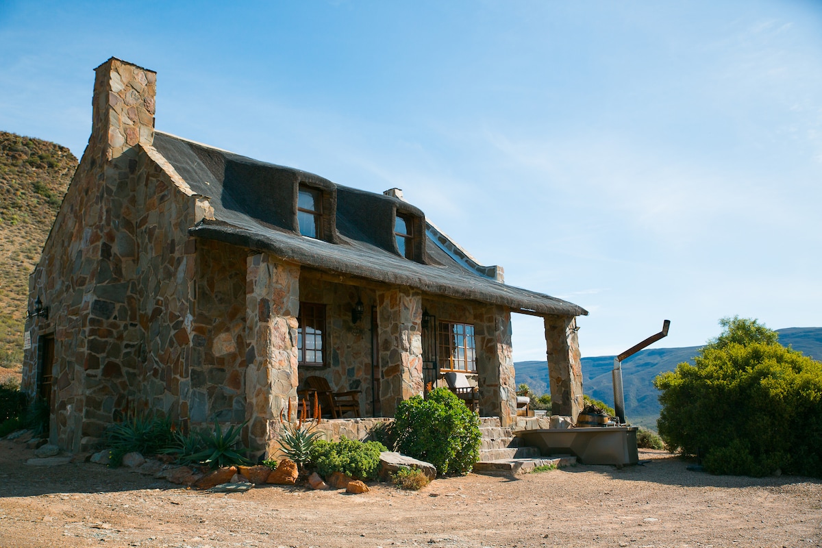 A stone cottage is set against a backdrop of rolling hills, displaying a pitched roof and a covered front porch. Natural stone walls add rustic charm, while surrounding greenery complements the earthy tones of the cottage. A barbecue area can be seen nearby.