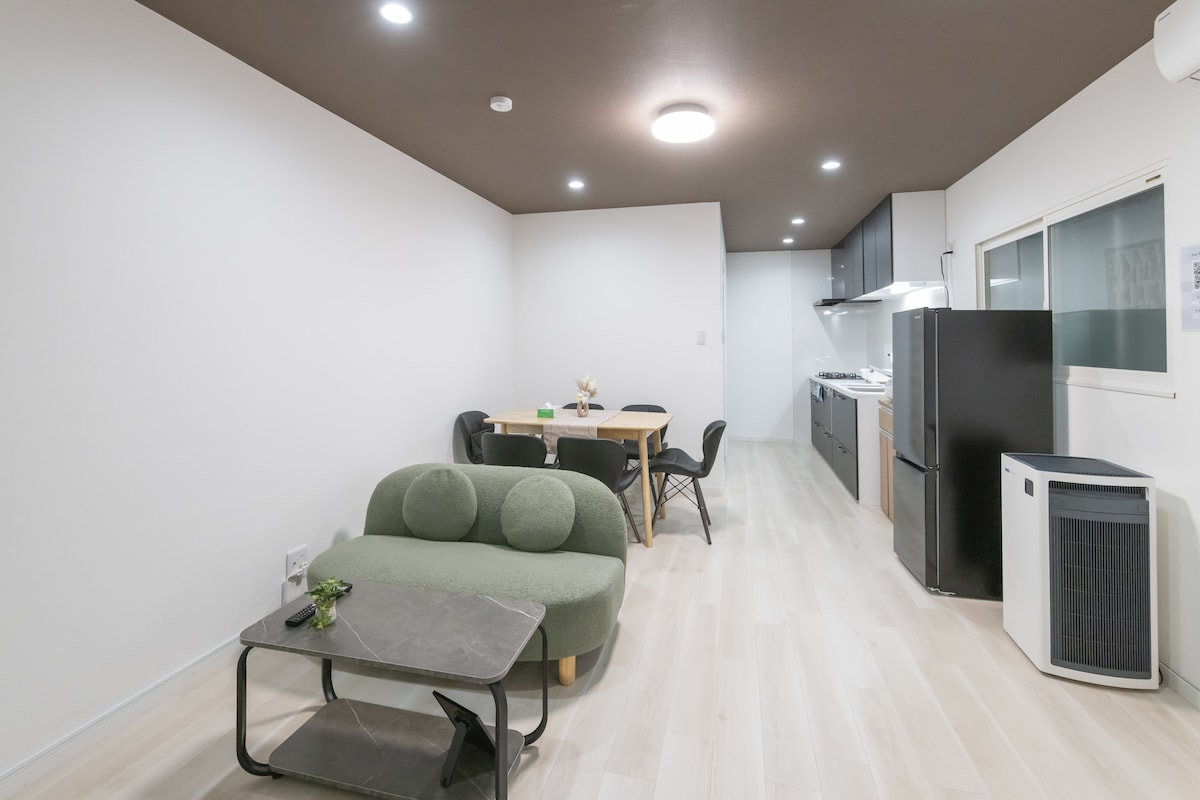 An open living space features a dining table with chairs and a cozy green sofa positioned around a low coffee table. The kitchen area, equipped with modern appliances, includes a refrigerator and cooking facilities, providing a functional and inviting atmosphere.