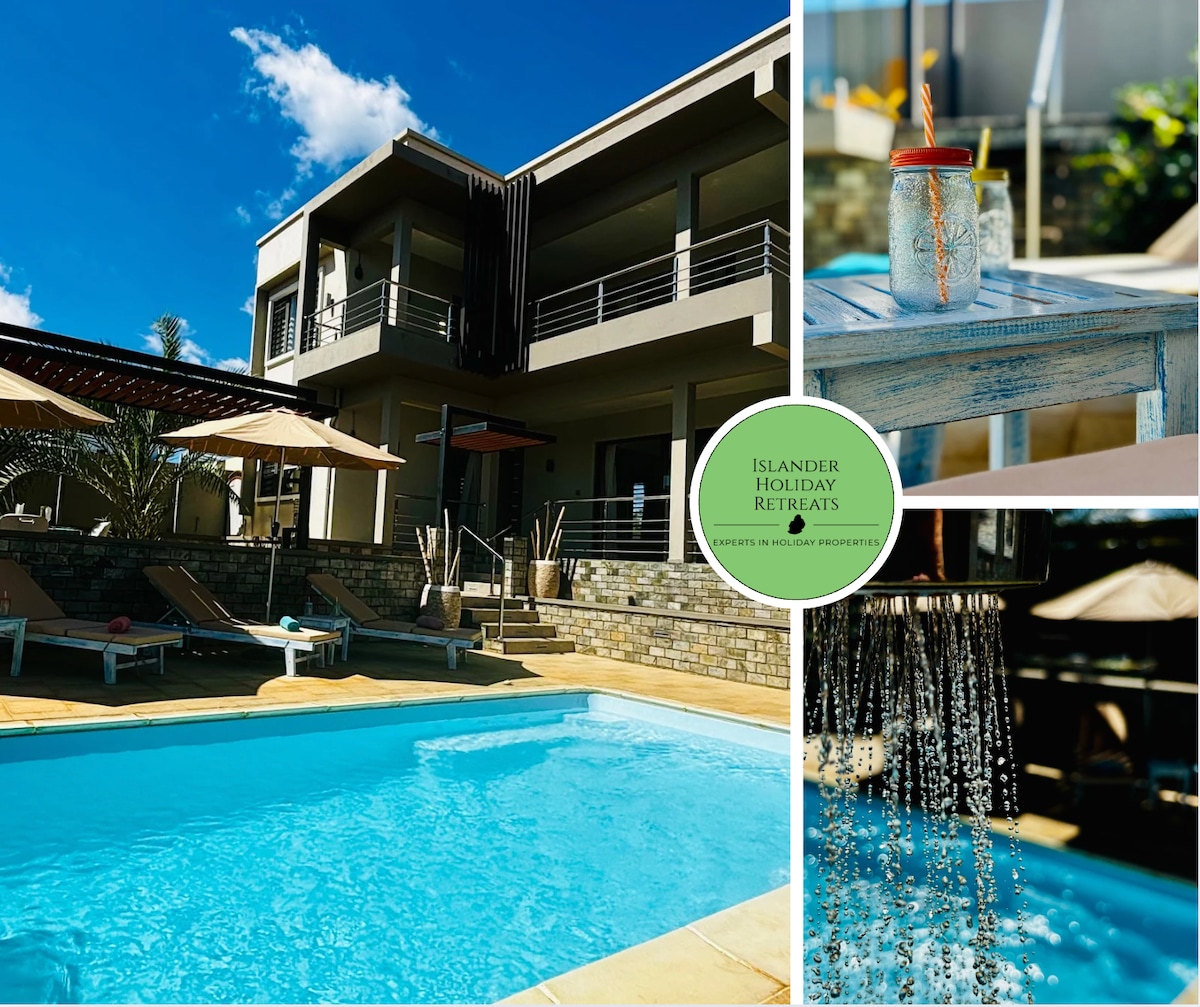 The outdoor area features a refreshing pool surrounded by sun loungers under shaded umbrellas. A patio area with a natural wood table and decorative accents adds to the relaxed atmosphere, while the modern villa's architecture provides an inviting backdrop.