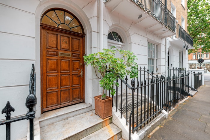 The entrance to the building is characterized by a large wooden door with decorative glass panels. A planter with greenery is positioned beside the steps, which are framed by a black wrought-iron railing. The façade features classic architectural details indicative of the area.
