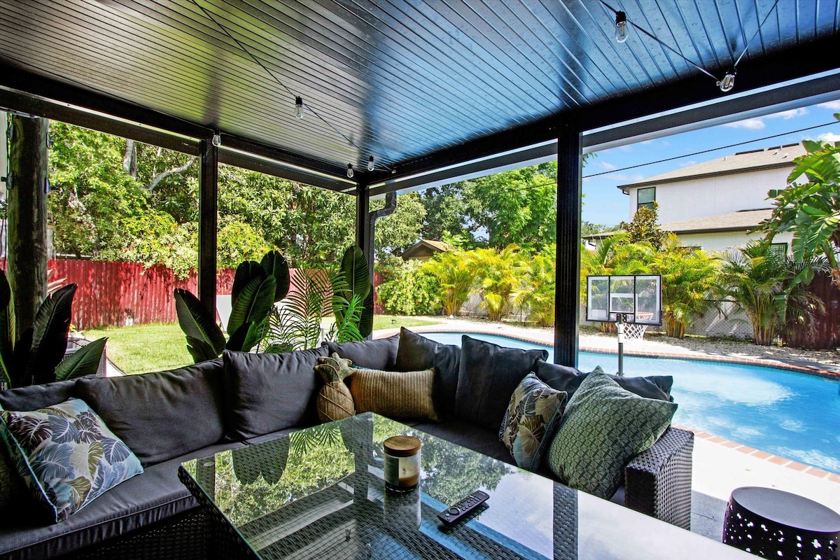 A spacious screened-in patio offers a comfortable sitting area featuring a large sectional sofa adorned with decorative pillows. A glass table occupies the center, with outdoor lighting above, and lush greenery visible through the screened walls, providing a view of the inviting pool area.