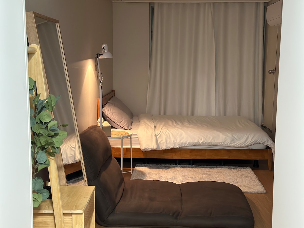 A compact living space features a spacious bed with a neatly arranged duvet and a bedside table with a lamp. A comfortable lounge chair is positioned nearby, with a large mirror reflecting natural light from the window draped in white curtains.