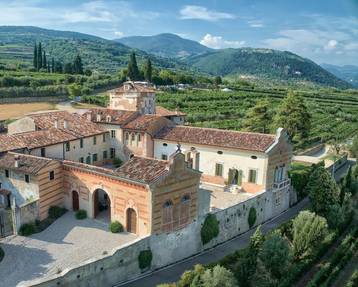 An elegant villa with a rustic facade is set against rolling hills and lush vineyards. The structure features a combination of brick and stone walls, framed by carefully landscaped greenery. A serene landscape stretches in the background, enhancing the tranquil atmosphere of the property.