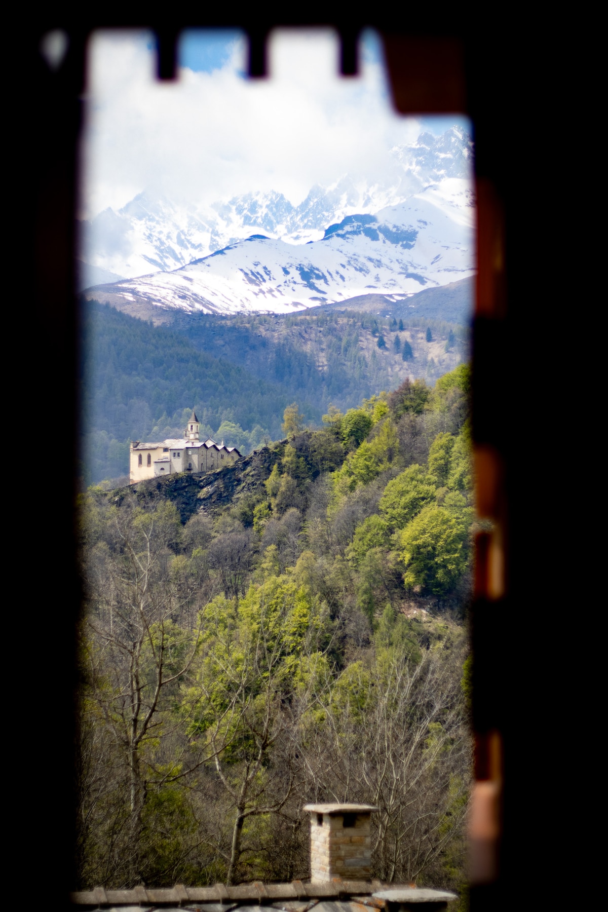 A view of a distant building perched on a hillside, surrounded by lush greenery. Majestic snow-capped mountains rise in the background, partially obscured by soft clouds. The scene is framed by a window’s edge, offering a glimpse of the tranquil landscape.