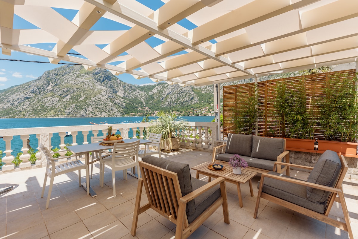 The spacious terrace features comfortable outdoor seating, including a sofa and chairs, arranged around a wooden coffee table. A dining area with a table is also present, offering unobstructed views of the sea and surrounding mountains through large openings in the overhead canopy.