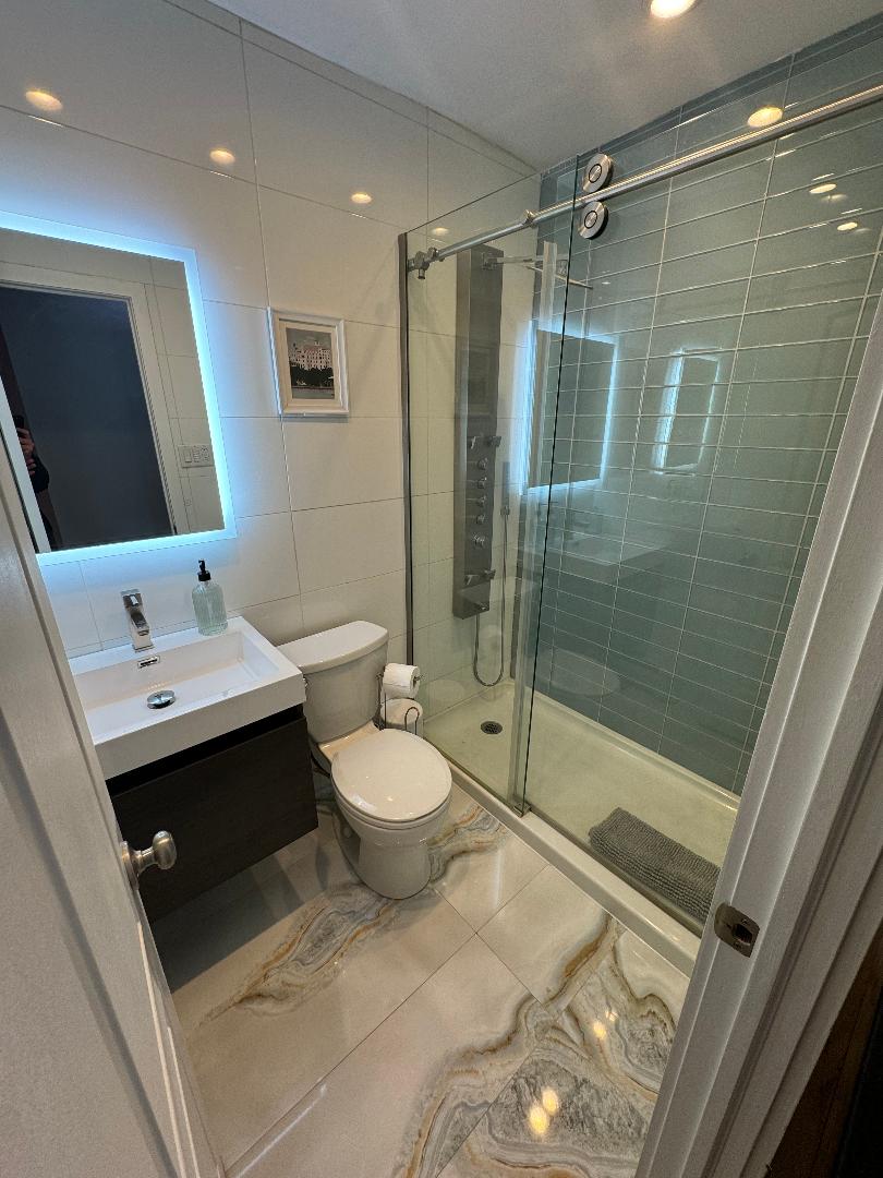 A modern bathroom is equipped with a glass shower featuring a sleek shower panel. White tiles and light-colored cabinetry enhance the clean aesthetic. A backlit mirror above the sink provides illumination, and the floor is covered in marble-patterned tiles, adding a touch of elegance.