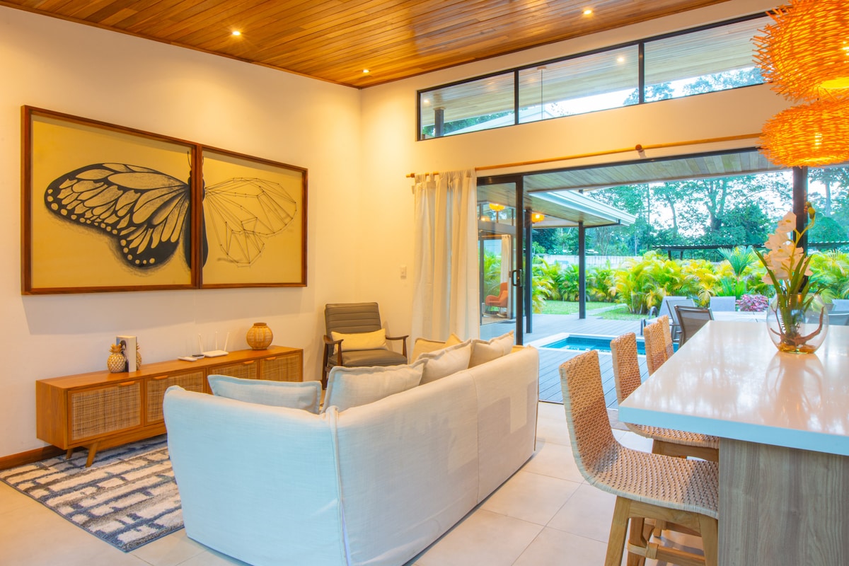 A bright living area features a large abstract artwork of a butterfly on the wall, accompanied by a comfortable white couch and a wooden sideboard. Large windows allow natural light to fill the space, revealing views of the outdoor garden and pool area.