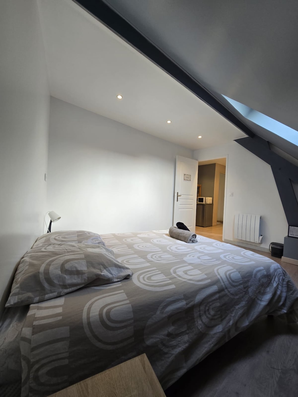 A private bedroom features a spacious bed dressed in a patterned duvet. Soft LED lights illuminate the room, highlighting the minimalistic decor with a small bedside lamp. A doorway leads to a well-lit bathroom, and natural light streams in through a skylight.