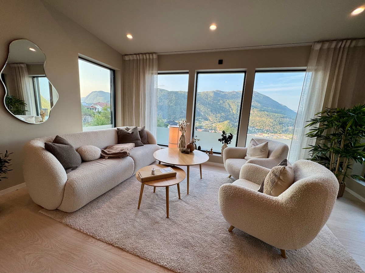 A cozy living area features large windows offering views of the surrounding mountains and valley. A round coffee table is positioned between a textured sofa and two armchairs. Soft textiles and greenery add an inviting touch to the space.