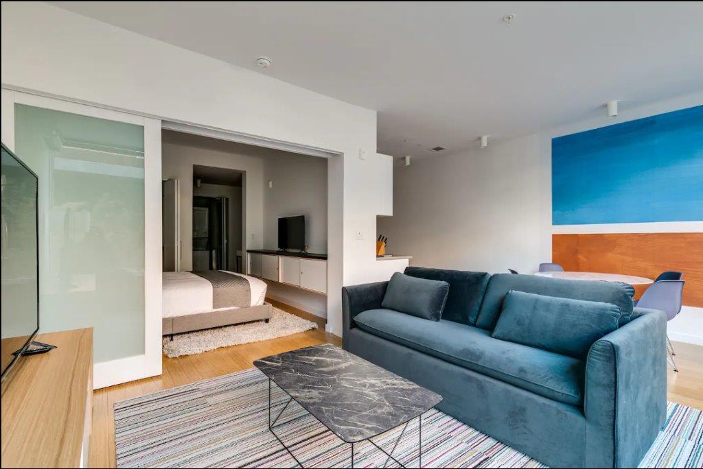 A spacious living area features a comfortable dark blue sofa and a coffee table with a gray marble top. A textured area rug lies beneath, and the open layout provides a view into the softly lit bedroom with a cozy bed, surrounded by light-colored walls.