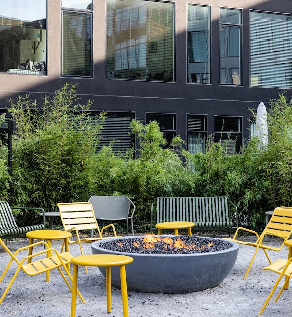 A circular fire pit with flames is surrounded by six modern chairs in a landscaped courtyard. Lush green bamboo plants provide a natural backdrop, and large windows from an adjacent building reflect the outdoor space.
