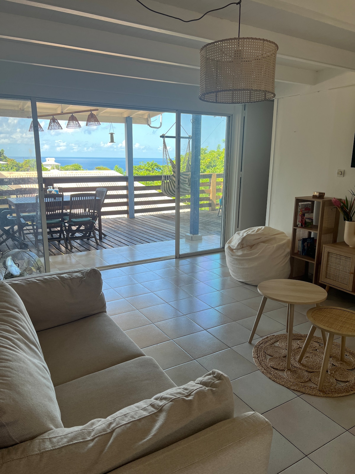 A comfortable living area features a light-colored sofa facing sliding glass doors that lead to a spacious terrace. A small round table set is positioned beside a woven rug. The outdoor dining area, visible through the glass, offers views of the Caribbean Sea.