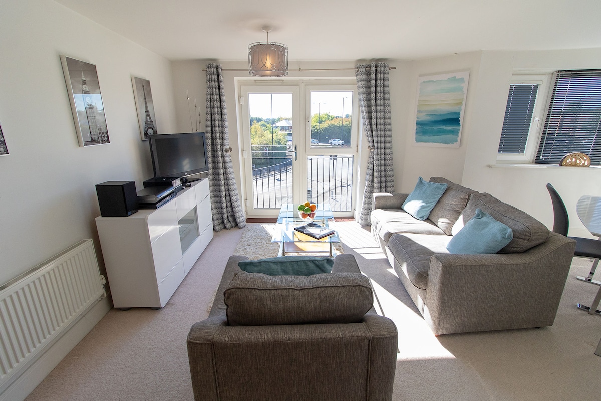 A spacious living area features two grey sofas adorned with light blue cushions. A glass coffee table sits centered on a cream rug. A smart TV is positioned on a white console, and large doors open onto a balcony, allowing natural light to fill the room.