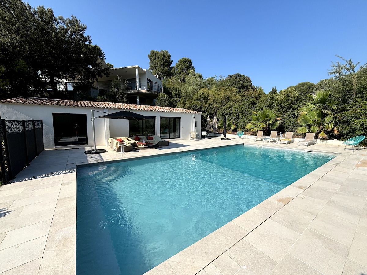 A spacious pool area is surrounded by a stone patio, featuring lounge chairs and umbrellas for relaxation. Lush greenery borders the space, providing a natural backdrop. The structure nearby offers large glass doors that invite views of the pool and garden.