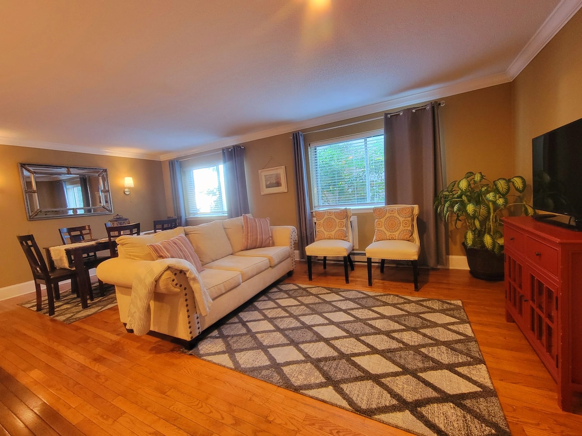 A spacious living area features a light-colored sofa accompanied by two patterned chairs. A dining table is positioned nearby, while large windows allow natural light to brighten the room. A vibrant plant enhances the welcoming atmosphere, and warm wooden floors complete the setting.