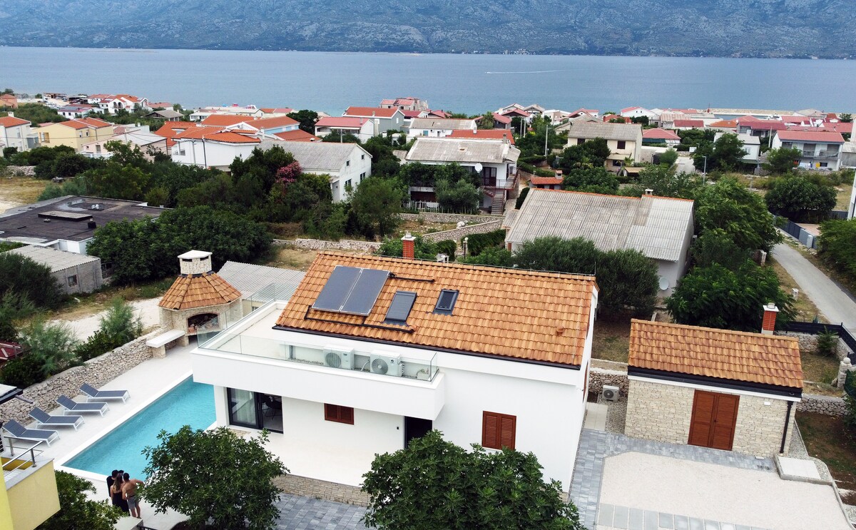 An aerial view of a modern house with a red-tiled roof and solar panels is presented. The property features a swimming pool surrounded by lounge chairs, while the backdrop includes the shimmering sea and distant mountains, providing a sense of tranquility and connection to nature.