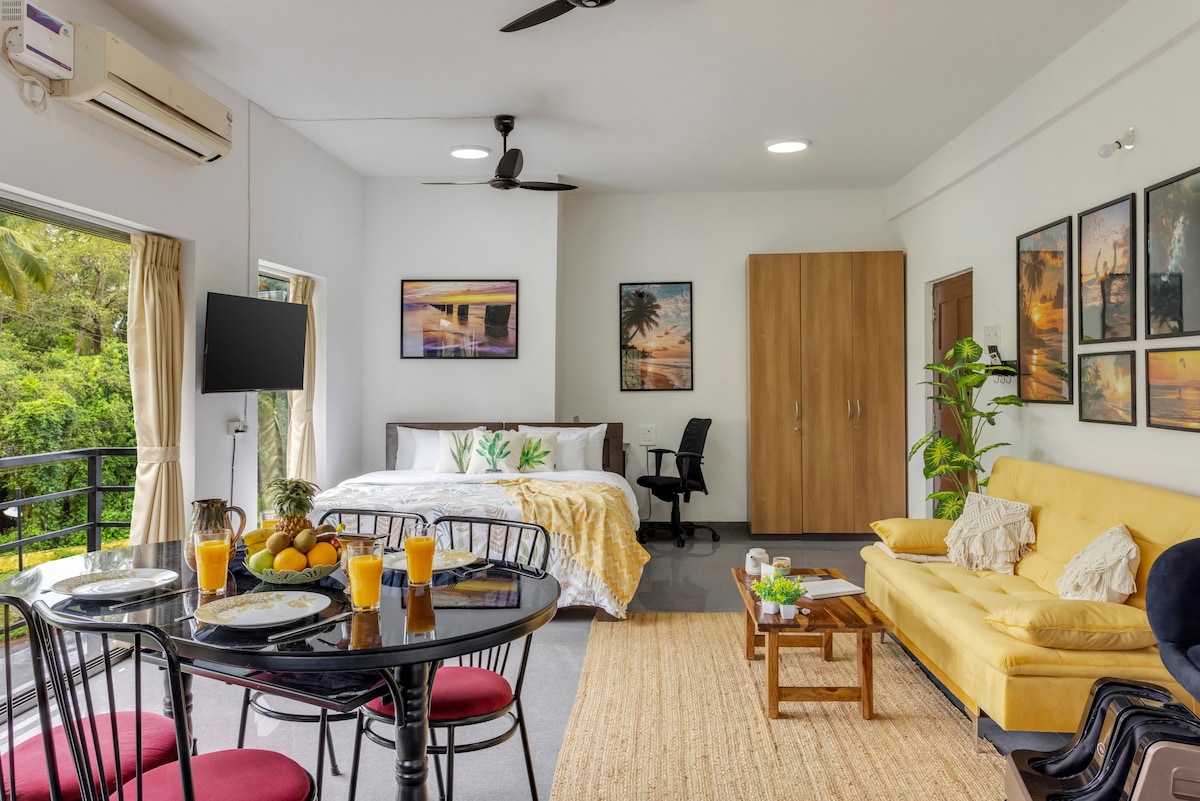 A bright and open living area includes a dining table set for four, featuring fresh fruit and drinks. A spacious bed is dressed in soft linens with artwork on the walls. A cozy seating area with a yellow sofa enhances the inviting environment.