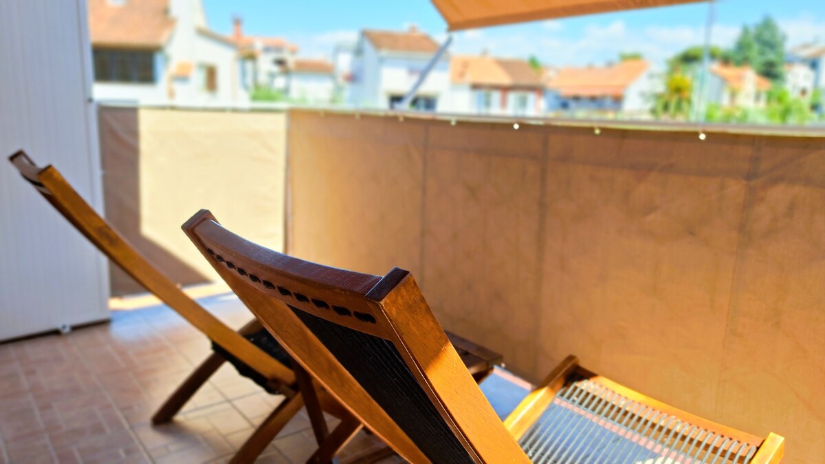 Two wooden lounge chairs are positioned on a balcony, providing a comfortable seating area. A sunshade extends overhead, partially blocking the sunlight. The background features a view of neighboring rooftops and greenery under a clear blue sky.