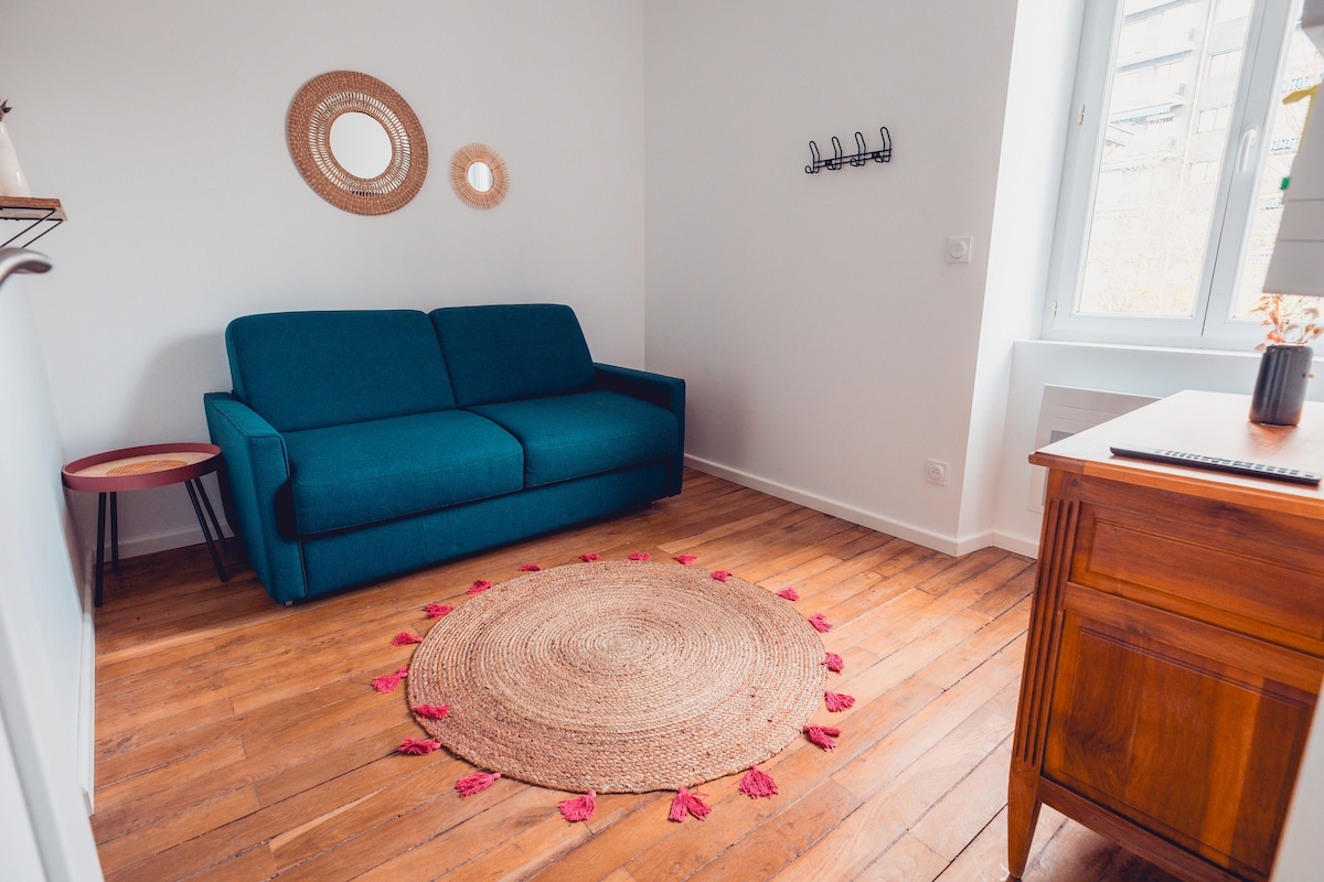 A bright seating area features a teal sofa positioned against a white wall. A round jute rug with pink tassels lies on the wooden floor. Natural light enters through a window adorned with simple coverings, while a small side table and a decorative mirror add subtle accents to the space.