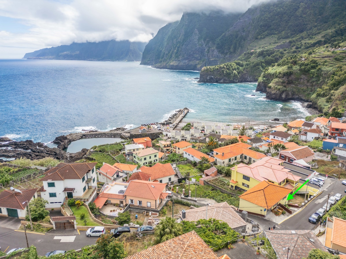 A panoramic view captures a coastal village with colorful rooftops, nestled against a rugged coastline. The azure waves gently lap at the shore, while lush green mountains rise in the background, partially veiled by clouds. A small harbor and beach can be seen in the distance.