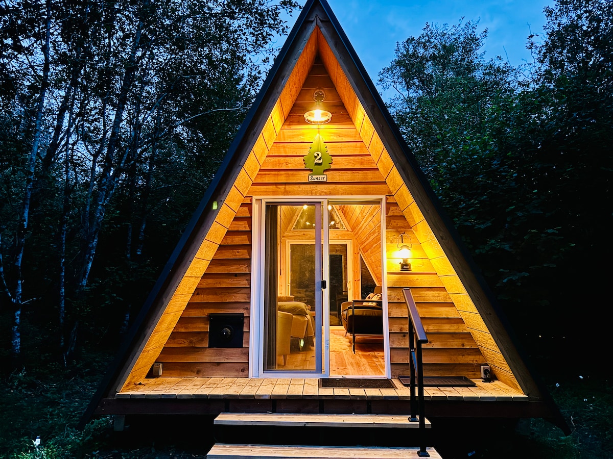 The A-frame cabin exterior is illuminated at dusk, showcasing its wooden facade and triangular design. A cozy porch with steps leads to glass doors, inviting entry. Soft lighting enhances the structure's architectural features, surrounded by lush greenery.