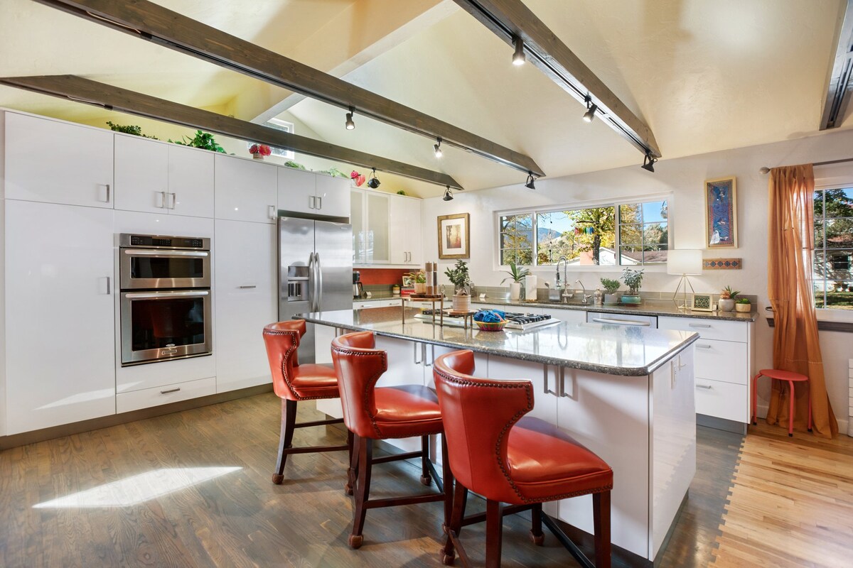 A modern kitchen is equipped with sleek white cabinetry and stainless-steel appliances. An island with a polished countertop is complemented by three red barstools. Large windows allow natural light to brighten the space, and decorative plants add a touch of greenery.