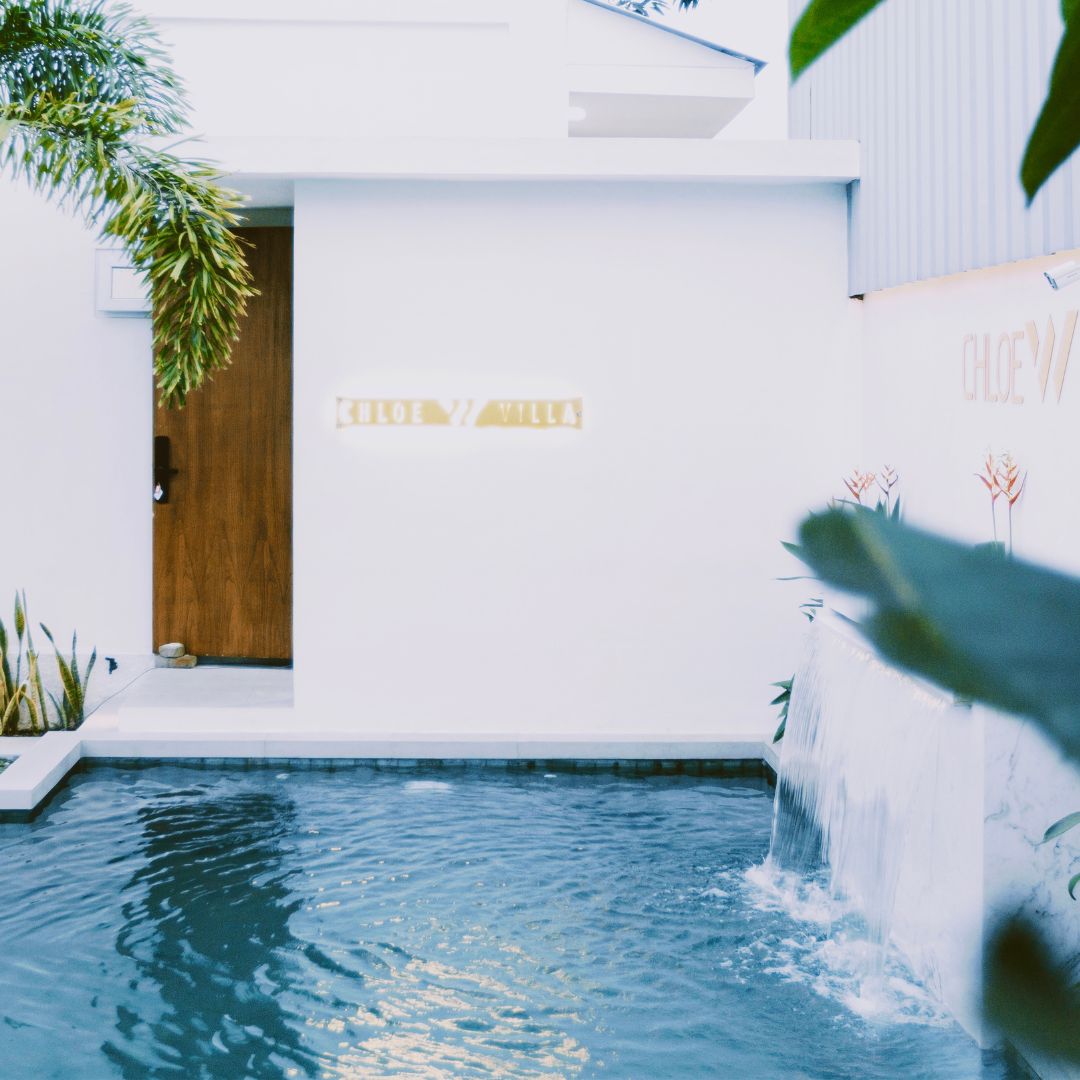 A serene entryway is captured, showcasing a modern facade with a wooden door. A gentle waterfall flows into a clear pool surrounded by tropical plants, enhancing the tranquil atmosphere. Soft lighting highlights the name 'Chloe W Villa' on the wall.