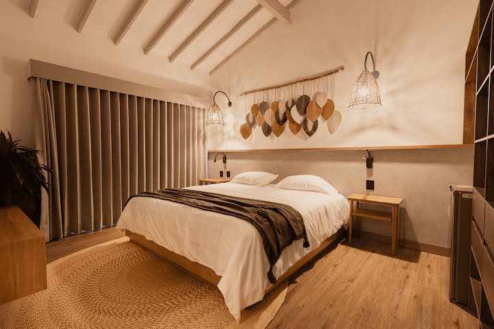 Pumzika katika chumba chenye utulivu chenye dari za juu, taa laini na mapambo yenye rangi ya udongo. Sanaa kubwa ya ukuta iliyosukwa na zulia la jute huongeza joto na muundo wa mbunifu. ️

