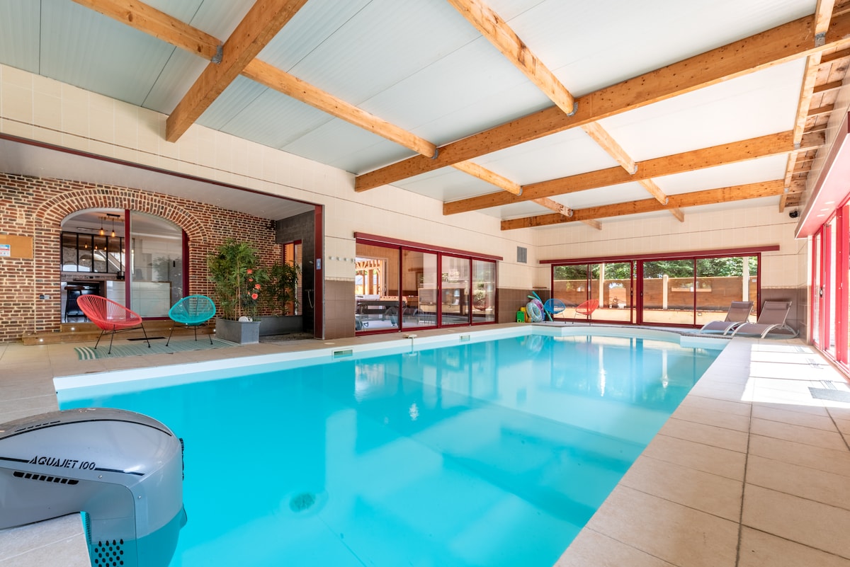 An indoor heated swimming pool is displayed, featuring a turquoise surface and surrounded by large windows with wooden beams overhead. Lounge chairs are arranged nearby, providing a space for relaxation. The atmosphere is illuminated by natural light entering from the expansive glass doors.