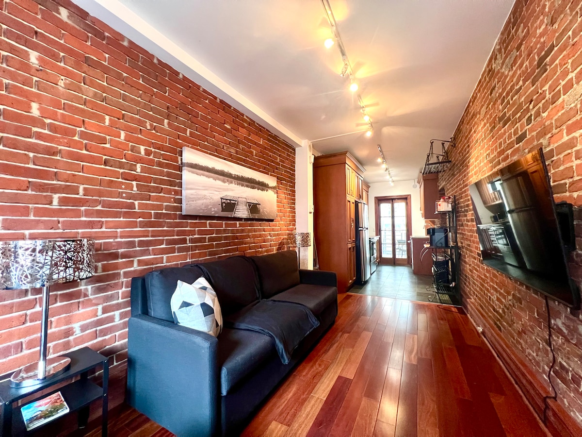 An inviting living area features exposed brick walls and hardwood flooring. A dark sofa provides seating, complemented by a small side table with a lamp. A wall-mounted TV is positioned opposite, and light fixtures illuminate the space, enhancing the cozy ambiance.