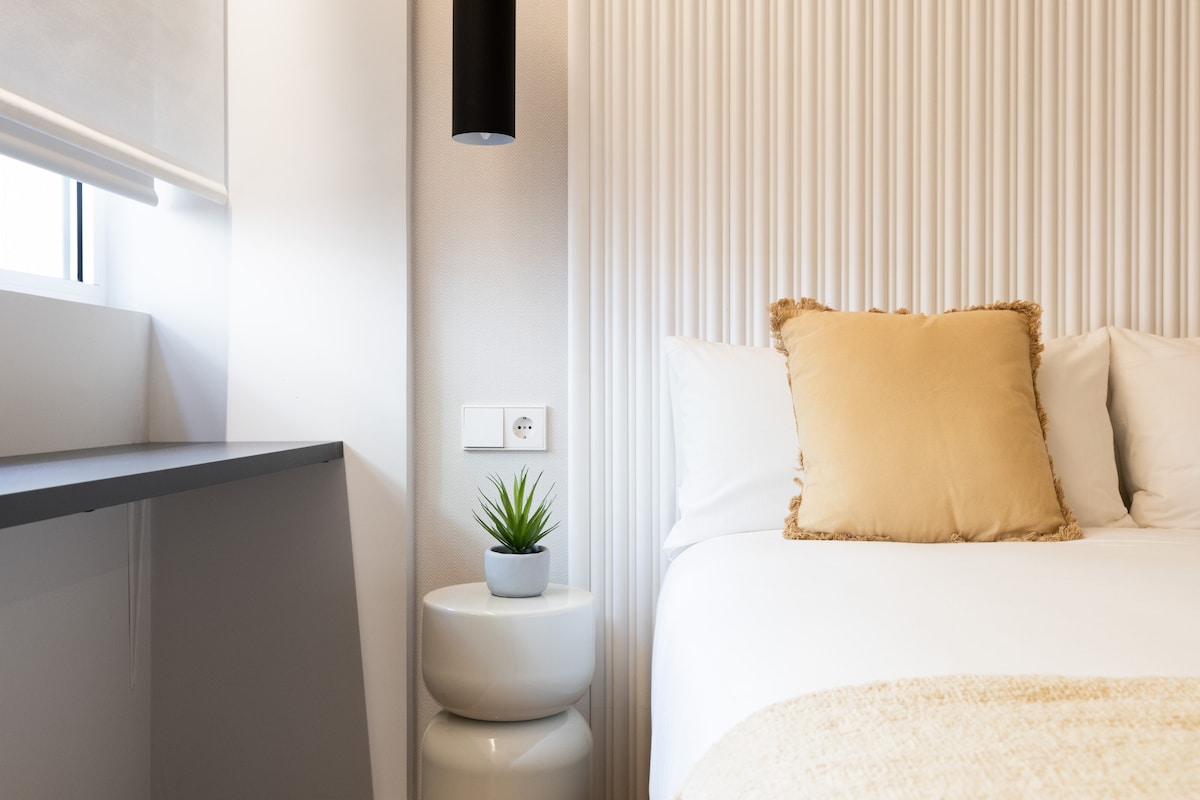 A cozy bedroom corner features a spacious bed with soft white linens and a decorative golden pillow. A small, white bedside table holds a potted plant. A black pendant light hangs above, complementing the textured accent wall.