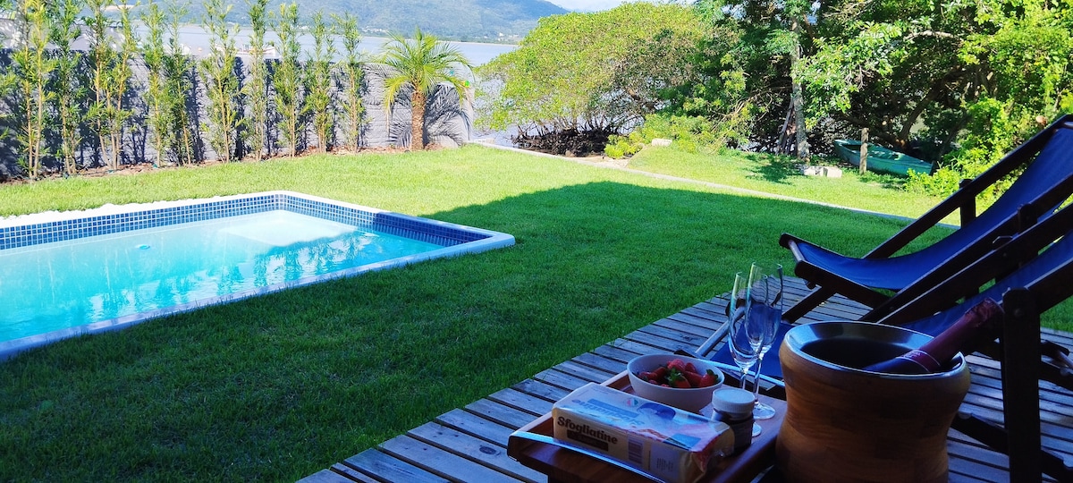 A tranquil outdoor scene is presented, highlighting a pool surrounded by green lawn and tropical foliage. Two loungers are positioned nearby, accompanied by a table set with a bowl of fruit and a bottle of champagne, overlooking a serene body of water.