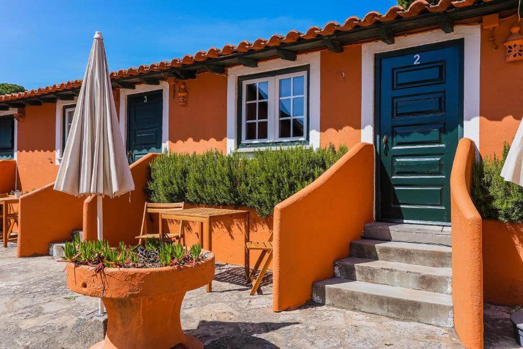 A welcoming exterior view of a vibrant orange building, showcasing two green entrance doors with a small staircase. A patio area is visible, featuring a wooden table and an umbrella, surrounded by landscaped greenery.