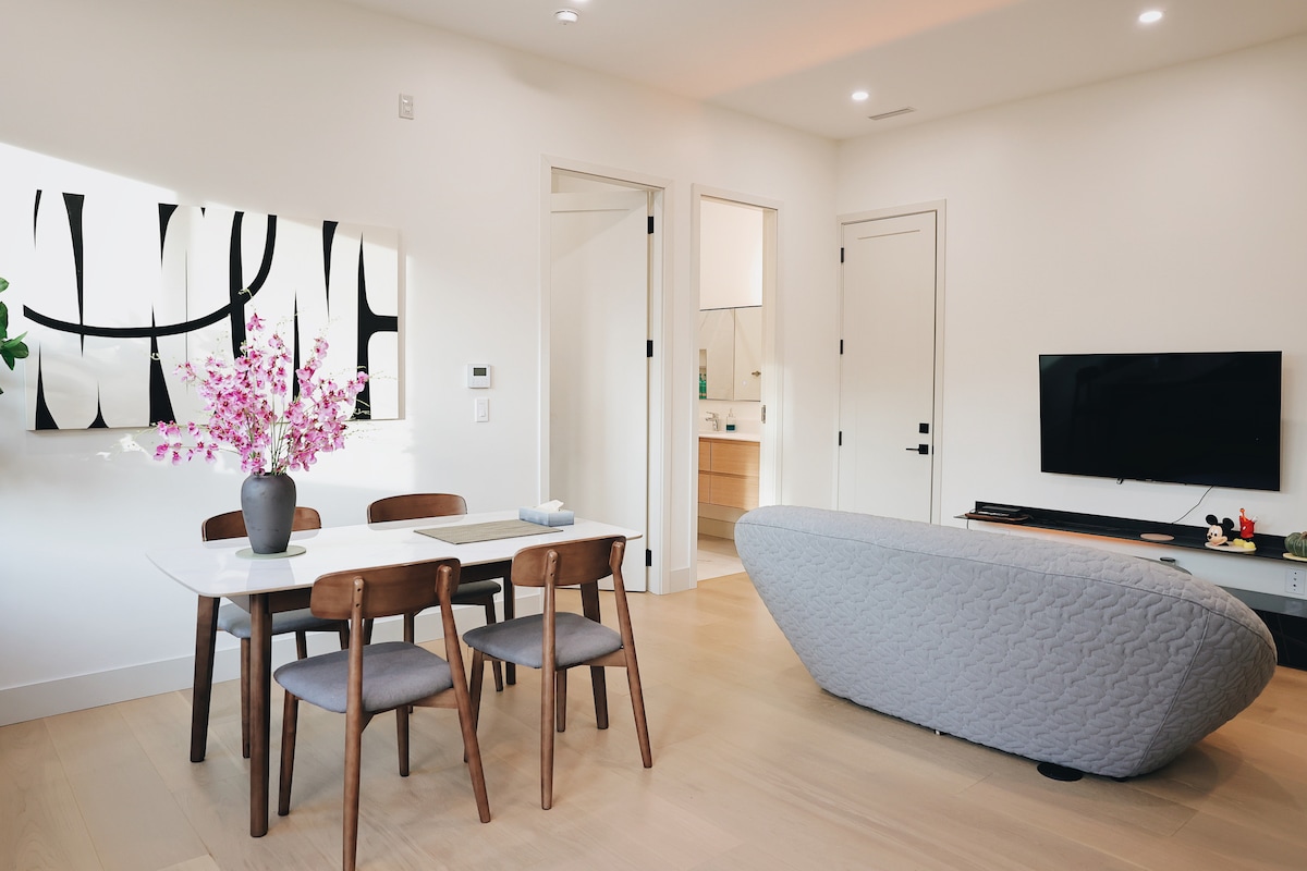 A modern dining area is presented with a sleek white table surrounded by four wooden chairs. A minimalist artwork adorns the wall, and a bouquet of flowers is placed in a vase on the table. A cozy couch is positioned nearby, enhancing the open layout.