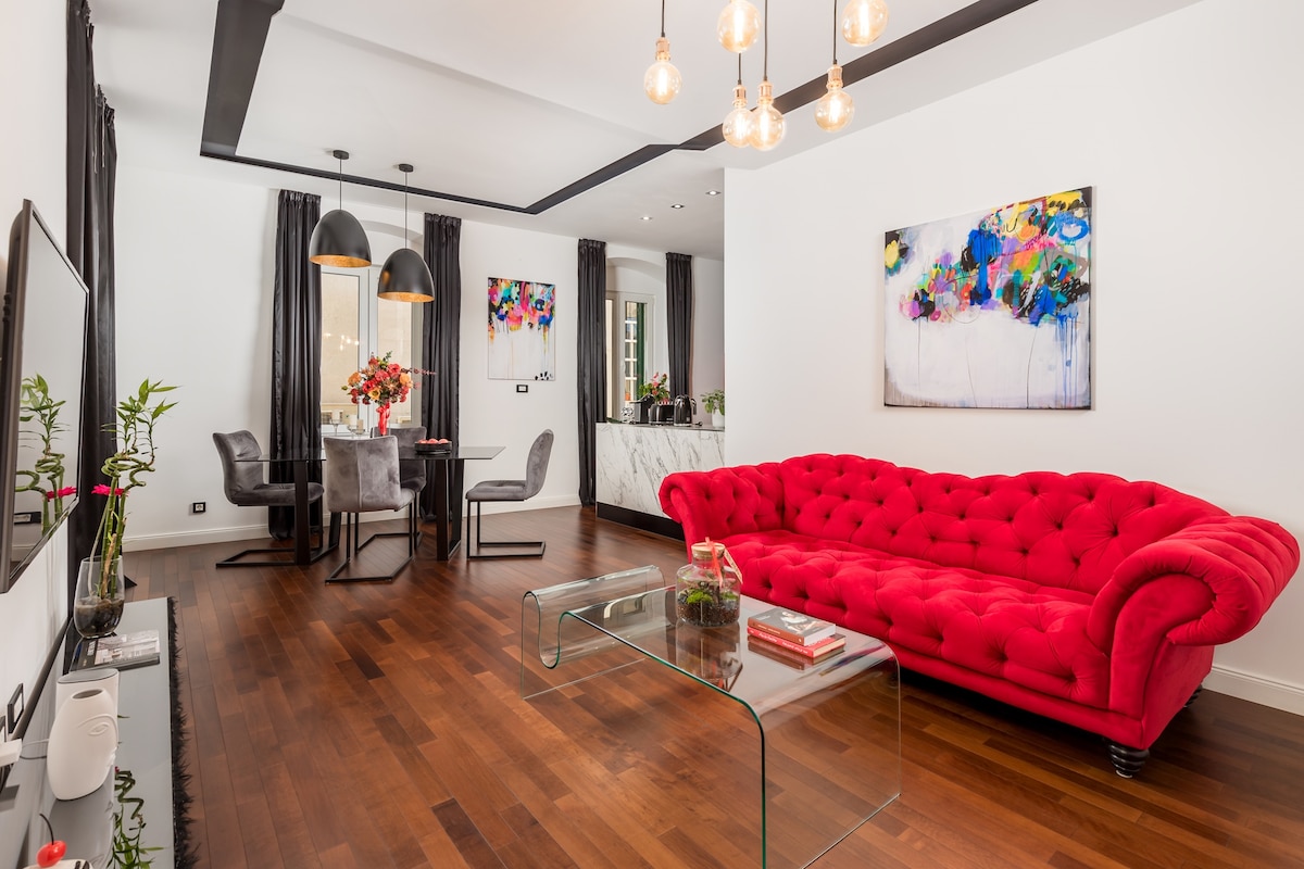 A spacious living room features a red tufted sofa and a glass coffee table. Bright artwork adorns the walls. Dark curtains frame large windows, allowing natural light to fill the room. A dining area is visible in the background, complimented by stylish chairs and minimalistic decor.